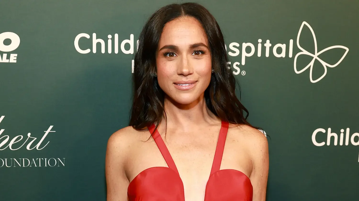 Meghan, Duchess of Sussex, wearing a red dress against a green background.