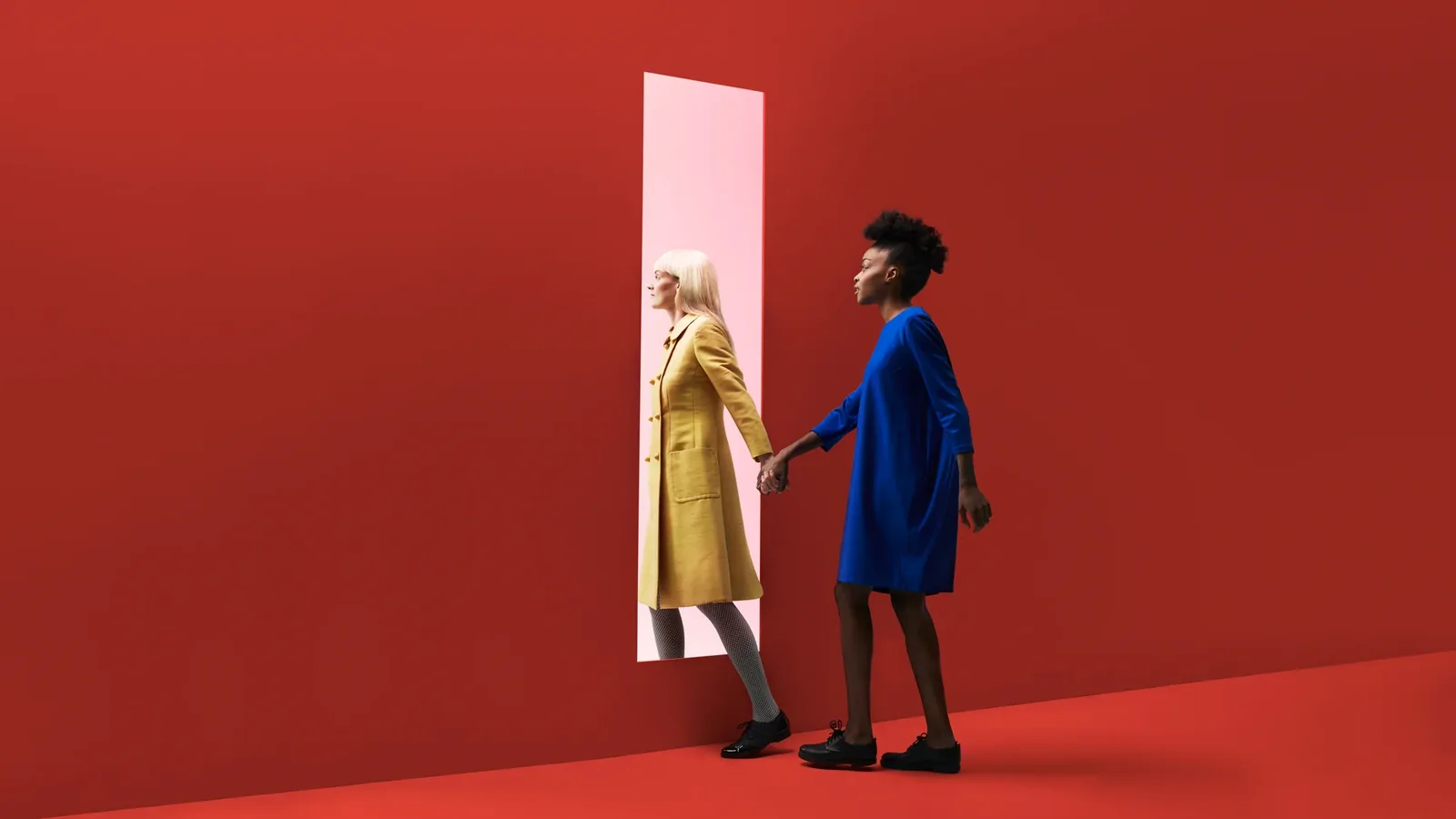 Two women holding hands as one steps through a tall opening in a red wall.