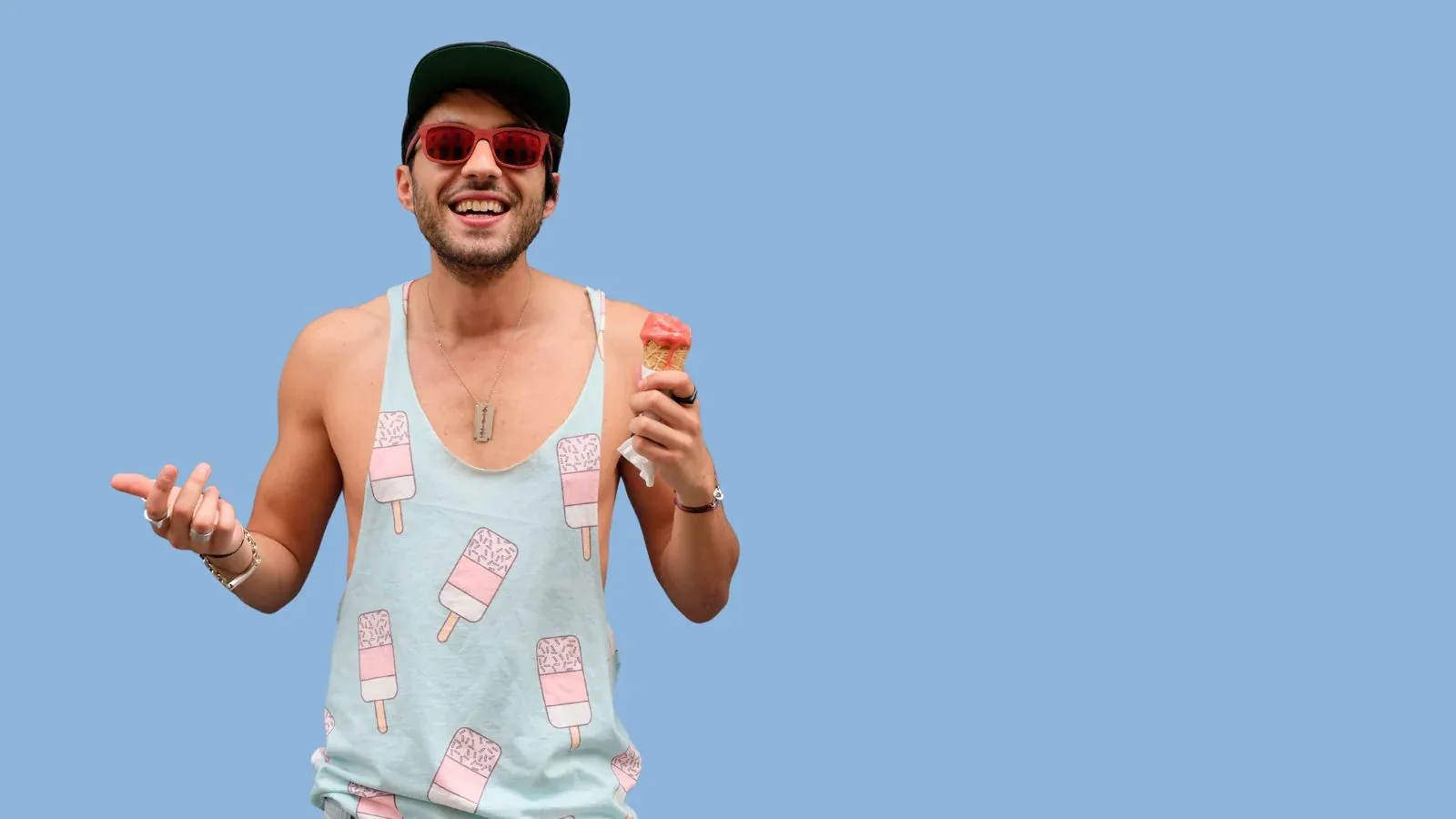 Smiling man in red sunglasses and an ice-lolly patterned vest holding an ice cream against a blue background.