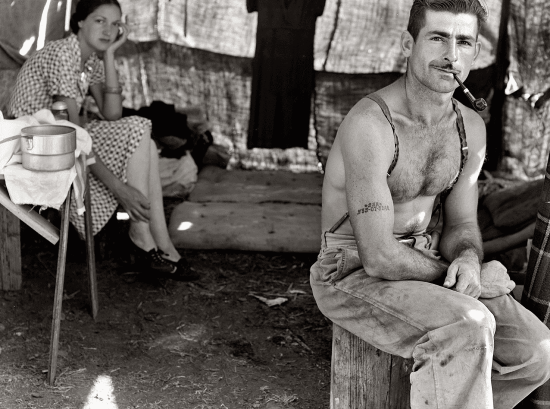 An unemployed lumber worker with a pipe sits in the foreground of a tent, with his wife seated behind him