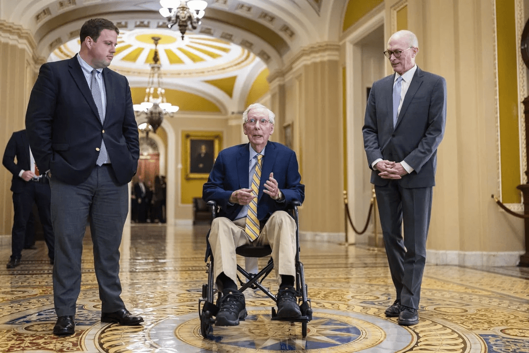 Mitch McConnell speaks from a wheelchair in a Capitol corridor with two men standing beside him